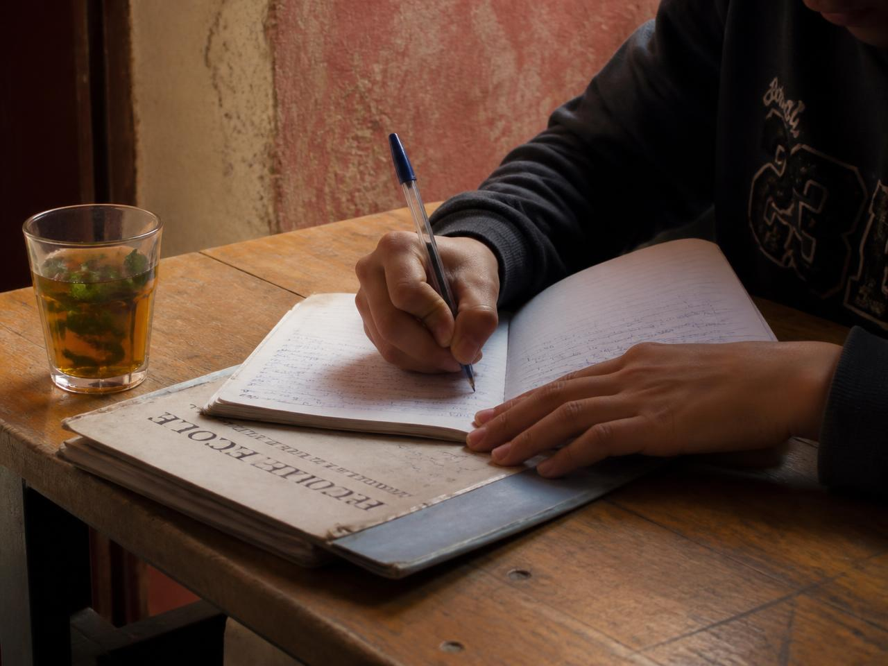 Jeune en train d’écrire dans un cahier, avec un verre de thé à la menthe, illustrant le pilier Scolarité, culture et ouverture